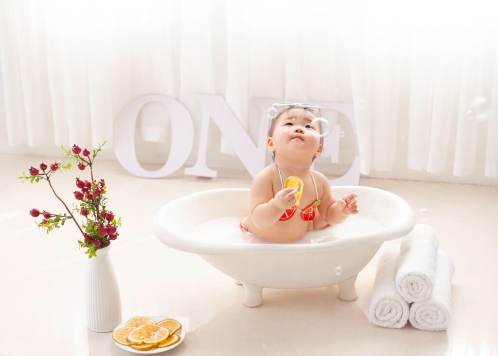 Cute Asian toddler in swimwear sitting in foamy water in bathtub and playing with soap bubbles