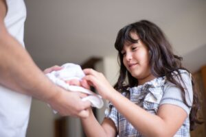 A young girl with brunette hair receives a face towel indoors with a smile.