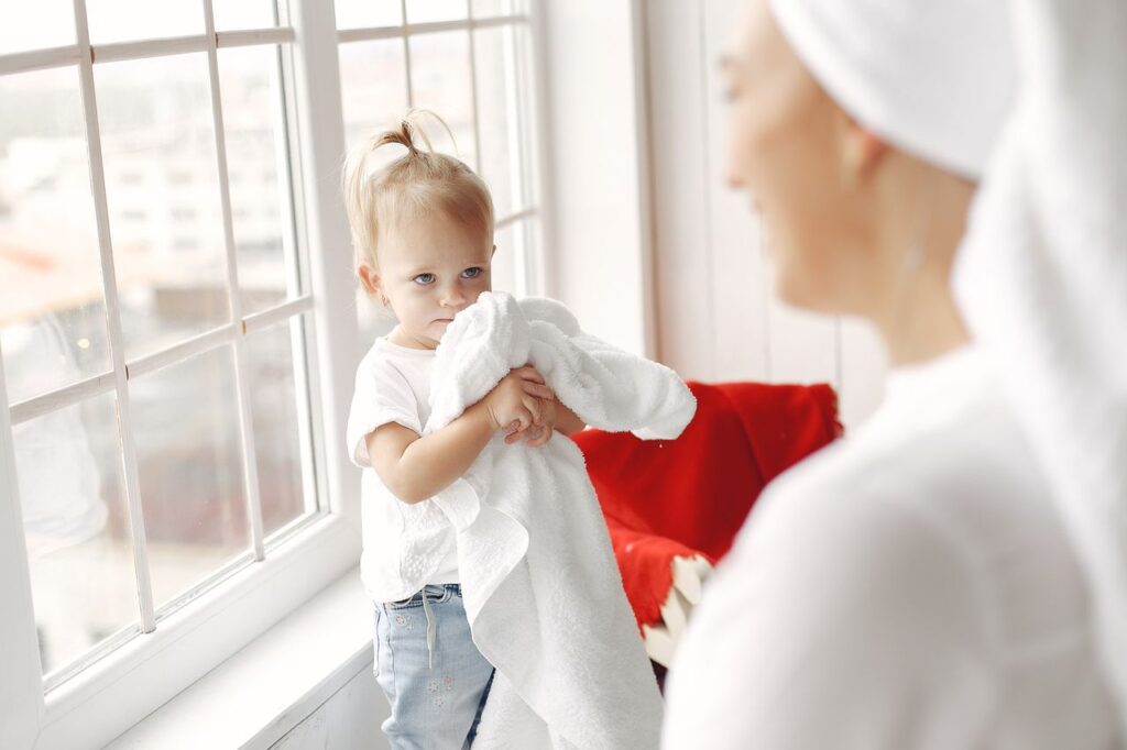 Adorable toddler holding towels by a window with mother nearby, creating a warm family moment.